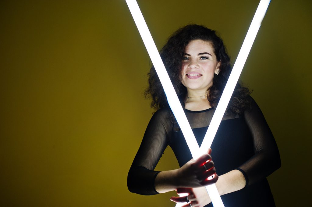 Curly brunette plus size model with two long led lamp against yellow wall.